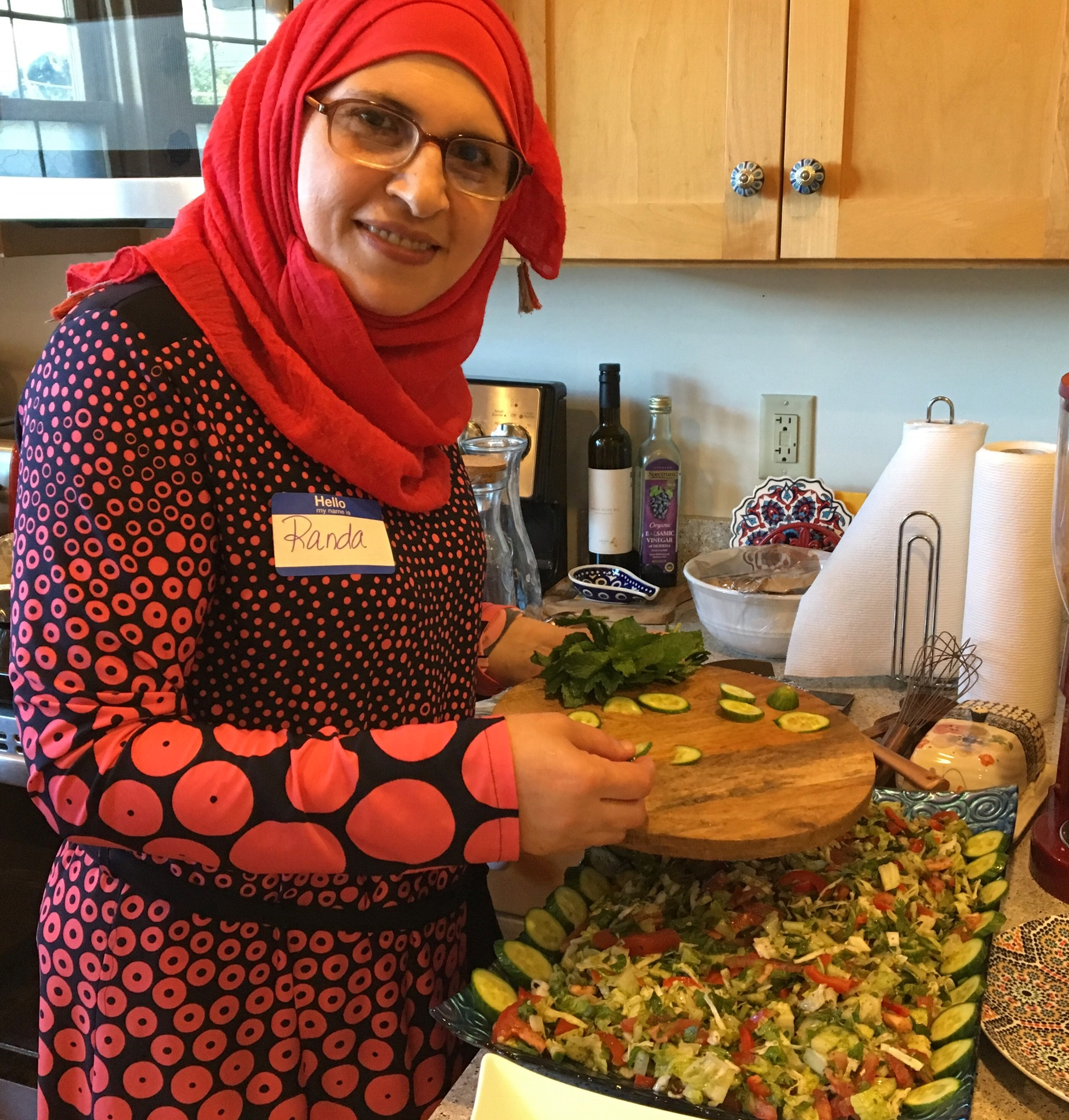 Randa of Sanctuary Kitchen and her Middle Eastern dip, mutabal. Chef Randa poses with ingredients, parsley, tomatoes and spices on a wood table.